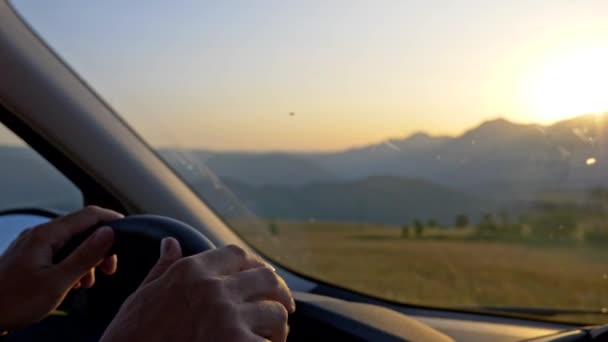 Dirigindo um carro a estrada rural durante o pôr do sol. Mãos masculinas segurando o volante. Prados e montanhas são vistos fora do carro. O sol está se pondo acima de uma montanha. UHD — Vídeo de Stock