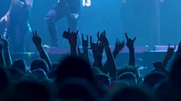 Pessoas numa multidão a saltar para o sulco durante um concerto de rock. Têm as mãos a mostrar o gesto dos chifres. O palco é iluminado com luz azul enquanto uma banda está se apresentando. 4K — Vídeo de Stock