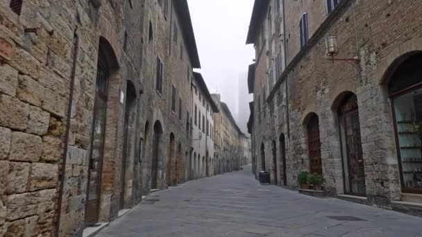 Caminhando pela rua estreita de San Gimignano, Itália coberto com nevoeiro pela manhã. Entradas para casas e lojas são vistas em ambos os lados da rua. Steadicam tiro, UHD — Vídeo de Stock