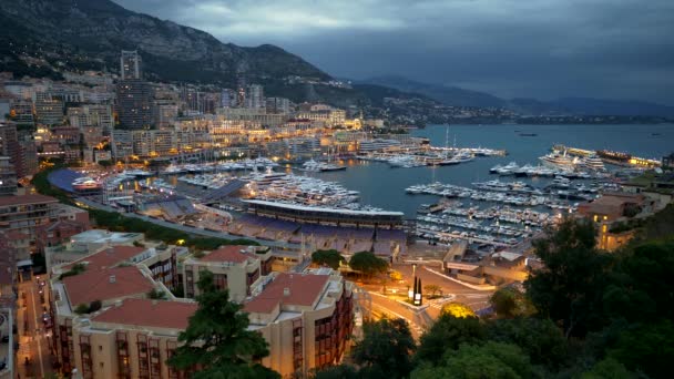 Crepúsculo em Mônaco. Carros estão indo as ruas, barcos e navios são atracados pela costa do Mar Mediterrâneo. Casas e ruas estão brilhando com luzes laranja da cidade. As montanhas estão acima da cidade — Vídeo de Stock