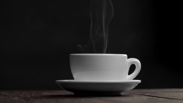 Hot white tea or coffee cup on a saucer against black background. Steam or smoke is coming from the cup. Slow motion shot — Stock Video