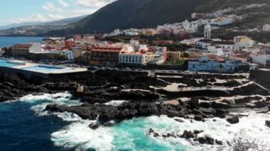 Garachico, Tenerife, İspanya. Güneşli bir günde adanın yanından uçmak. Dalgalar kıyıya vuruyor. Arabalar trafiğe giriyor. Hava görüntüsü, UHD