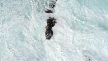 Fırtınalı okyanus dalgaları siyah kayalara çarpıyor ve bir sürü beyaz köpük ve su fışkırtıyor. Havadan yukarıdan aşağısı, UHD