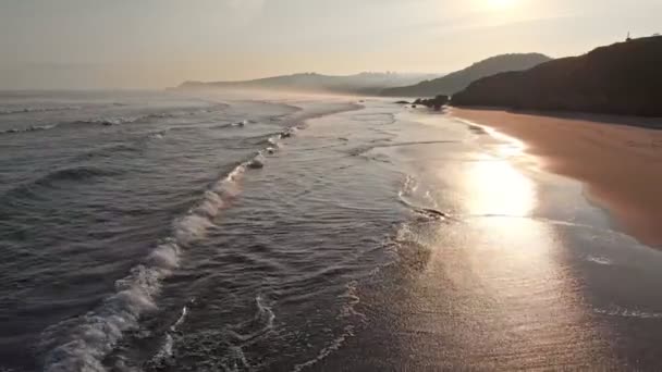 Spanyol Utara. Pantai pagi saat matahari terbit. Gelombang bergulir di pantai. Terang matahari memantulkan pasir basah. Kamera bergerak mundur. Tembakan udara, UHD — Stok Video