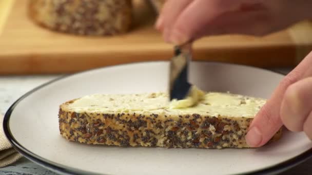 Przesuwne UHD strzał rozprzestrzeniania masła na chleb rzemieślniczy. Kobieta rozmazuje żółte masło na kromce chleba z nasionami sezamu — Wideo stockowe