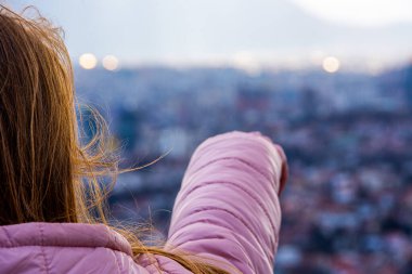 Kahverengi saçlı ve pembe ceketli kız şehre doğru uzaklığı gösteriyor.