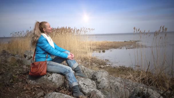 Die junge Frau sitzt auf Steinen am Ufer des Golfs und blickt auf das Wasser — Stockvideo