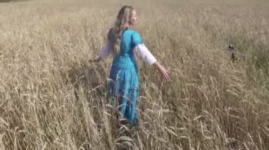 Uzun sarı saçlar olan ince kadın olgun buğday tarlada güneşli gün gider.