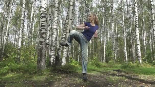 La mujer joven delgada con el cabello largo y delgado en la camiseta y los vaqueros lucha contra las artes marciales en el claro del bosque, la cámara lenta — Vídeo de stock