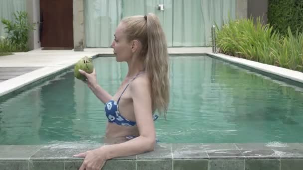 Die junge schlanke Frau im Bikini trinkt Kokosmilch aus einer Kokosmilch am Rande des Pools im tropischen Resort — Stockvideo