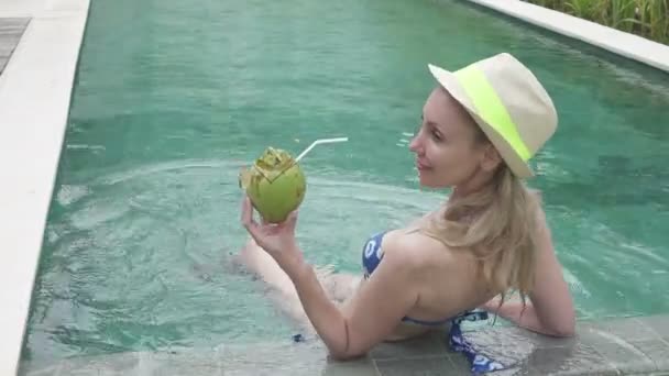 Die junge schlanke Frau im Bikini trinkt Kokosmilch aus einer Kokosmilch am Rande des Pools im tropischen Resort — Stockvideo