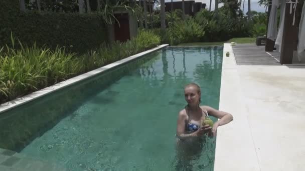 A jovem mulher esbelta com cabelo longo tem um resto na piscina e bebe coquetel de coco no resort tropical — Vídeo de Stock