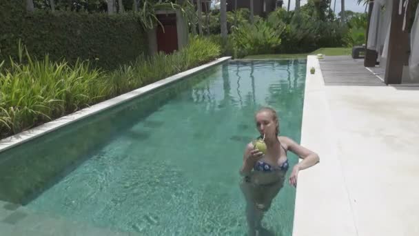 Молодая стройная женщина с длинными волосами отдыхает в бассейне и пьет коктейль из кокоса в тропическом курорте — стоковое видео