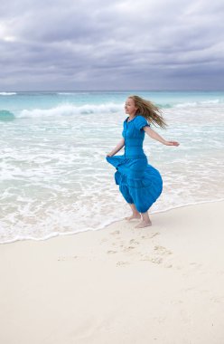 Kumlu bir sahilde, fırtınalı bir denizin kıyısında mavi bir elbiseyle rüzgarda dalgalanan ince saçlı genç bir kadın. (Küba, Varadero