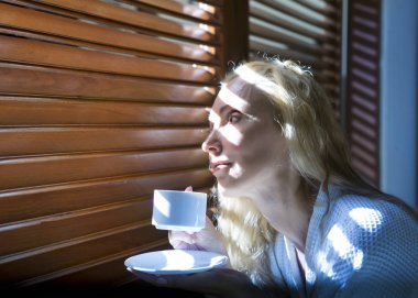 Kadın bornozlu ve elinde bir fincan kahveyle güneşlik penceresinin yanında çizgili bir kalça içinde.