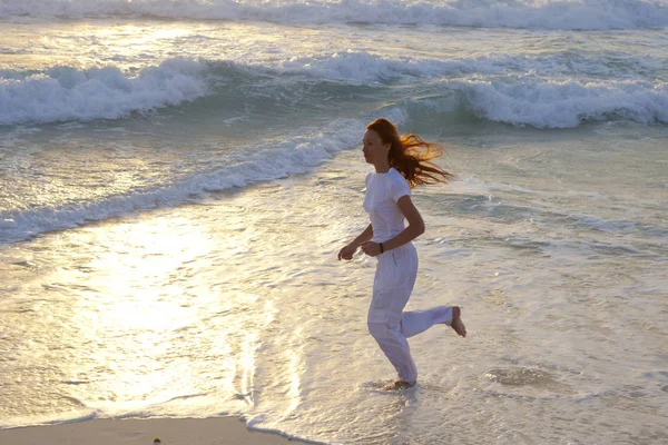 身穿白色运动服的纤细女子在沙滩上 在日出的背景下 沿着海浪的边缘晨跑 — 图库照片