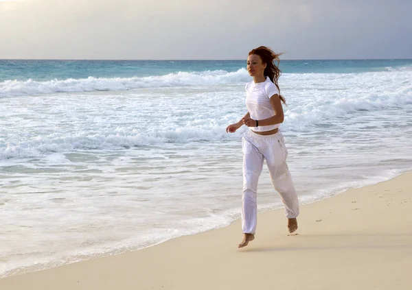 穿着白色运动服的纤细女子在沙滩上沿着海浪的边缘慢跑 — 图库照片