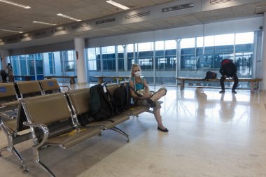 Coronavirüs salgını sırasında boş bir havaalanı salonunda tıbbi koruyucu maskeli bir kadın. Koltuklarda haç işaretleri var, yakın oturmayın, uzak durun..