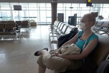 Tıbbi koruyucu maskeli kadın, Coronavirüs salgını sırasında boş bir havaalanı salonunda oturuyor..