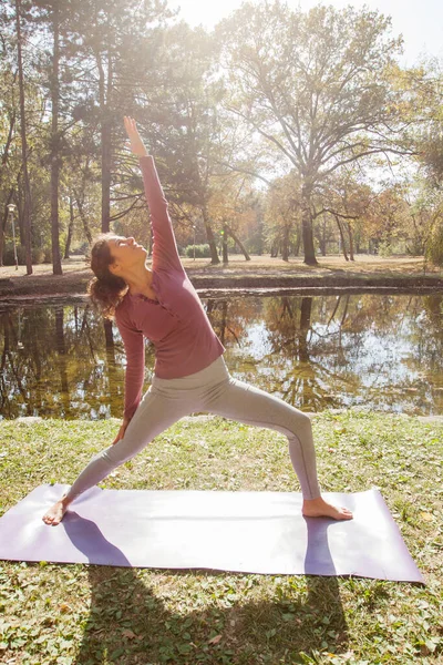 美丽的秋日清晨 年轻女子在湖边公园的垫子上练习瑜伽 — 图库照片