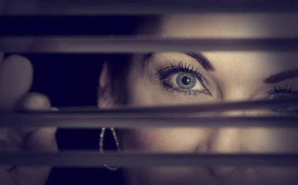 Retrato de uma mulher em pé na escuridão olhando através de persianas — Fotografia de Stock