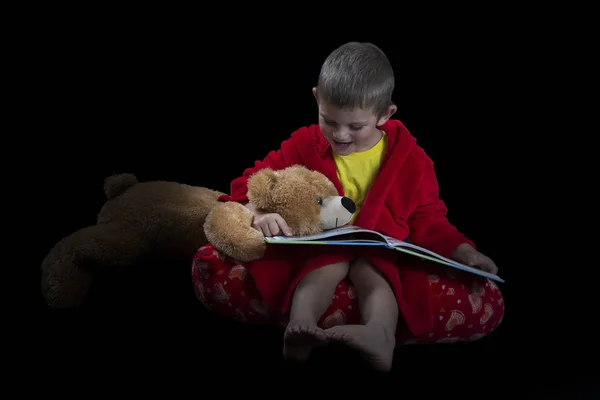 Смішний хлопчик з плюшевим ведмедем читає книгу перед сном — стокове фото