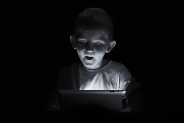 Funny boy sitting with a tablet in darkness before bed time in a