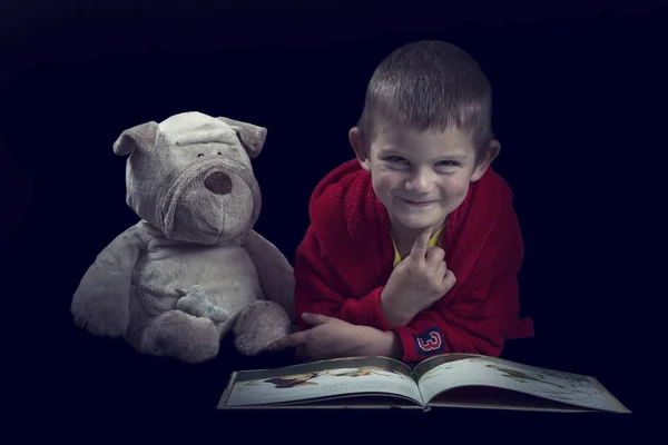 Funny boy with a stuffed dog reading a book for bed time in arti — Stock Photo, Image