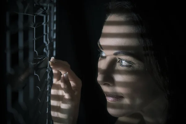 Portrait of a woman standing in darkness looking through blinds — Stock Photo, Image