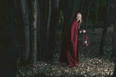 Attractive woman dressed a little red riding-hood walk in a dark forest with a lantern clipart
