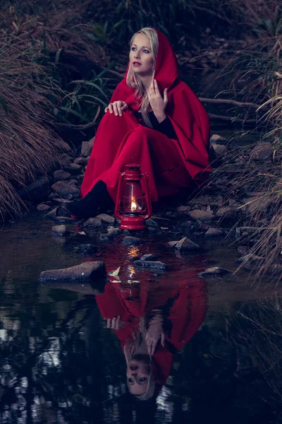 魅力的な女性は少し赤いライディングを着て ランタンで暗い森の中のストリームを横断フード — ストック写真