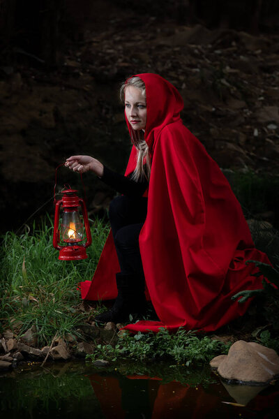 Attractive woman dressed a little red riding-hood cross a stream in a dark forest with a lantern