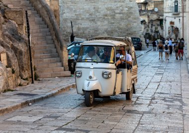 Matera, İtalya - 15 Eylül 2019: Matera şehrinde tarihi bir bölge olan Sassi di Matera 'da bir turist taksisi olarak kullanılan Piaggio Ape Calessino üzerinde turistler, antik mağara evleri ile tanınırlar. Basilicata. İtalya
