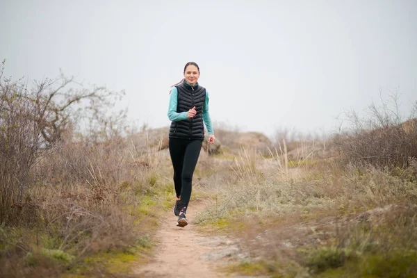 Krásná usměvavá žena běžící po horské stezce na chladném podzimním večeru. Sport a aktivní životní styl. — Stock fotografie