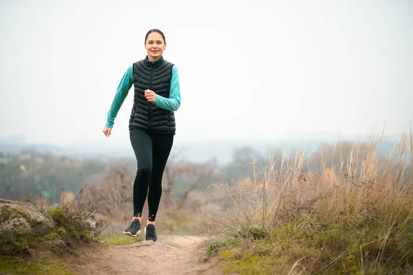 Hermosa mujer sonriente corriendo en el sendero de la montaña en la fría noche de otoño. Deporte y estilo de vida activo . — Foto de Stock