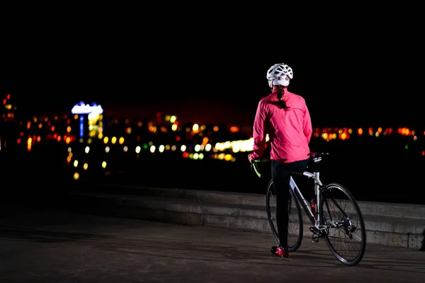 Женщина-велосипедистка отдыхает на велосипеде и смотрит на огни ночного города. Концепция здорового образа жизни и городского спорта — стоковое фото