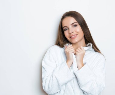 Gri arkaplanda poz veren uzun kahverengi saçlı, beyaz bornoz giyen güzel genç bir kadın.