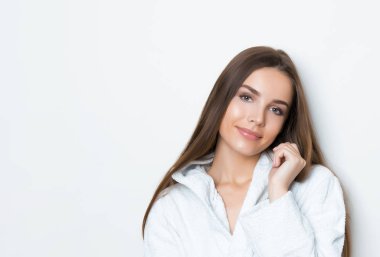Gri arkaplanda poz veren uzun kahverengi saçlı, beyaz bornoz giyen güzel genç bir kadın.