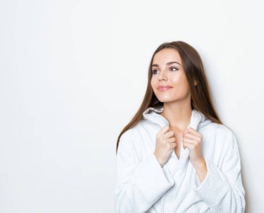 Gri arkaplanda poz veren uzun kahverengi saçlı, beyaz bornoz giyen güzel genç bir kadın.