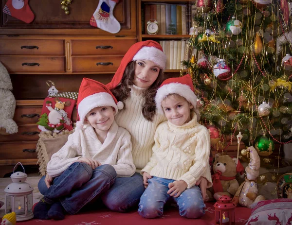 Kinderen zitten met moeder onder kerstboom in hoeden — Stockfoto