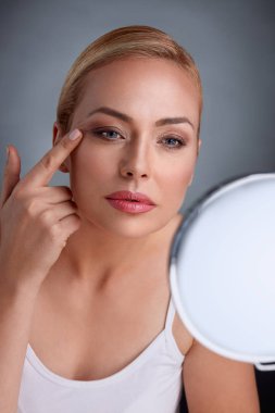 Beautiful woman looking her wrinkles in mirror clipart