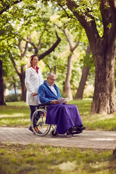 Infirmière poussant l'homme âgé en fauteuil roulant tout en lisant le livre — Photo