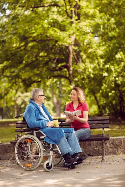 Junge Pflegerin liest Buch im Park behinderter Mann im Rad — Stockfoto