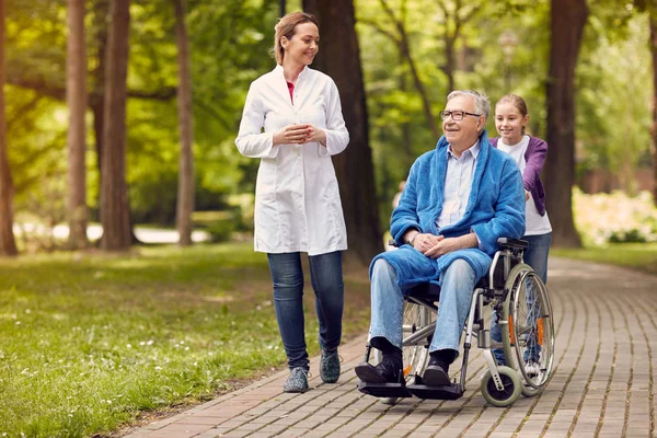 Älterer Mann im Rollstuhl mit Krankenschwester und Enkelin im Freien — Stockfoto