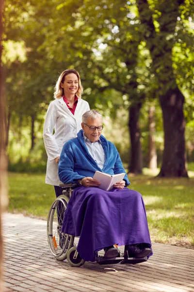 Infirmière souriante poussant l'homme âgé en fauteuil roulant tout en lisant bok — Photo