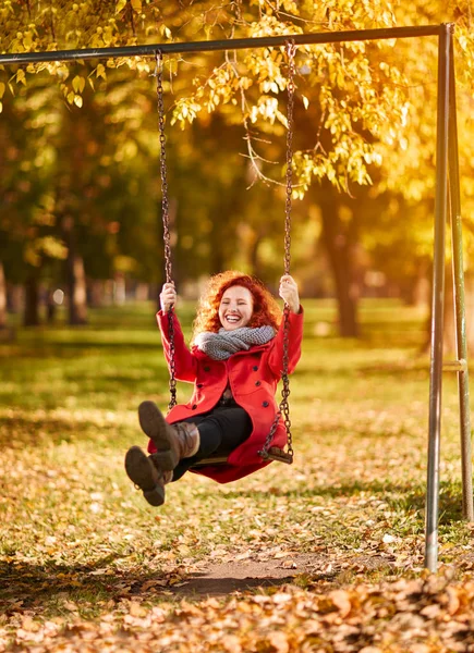 Счастливая рыжая женщина на качелях в парке — стоковое фото