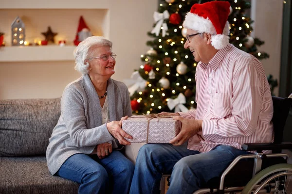 Ανώτερος άνθρωπος σε αναπηρικό καροτσάκι και χαμογελαστή γυναίκα με Χριστουγεννιάτικο δώρο — Φωτογραφία Αρχείου