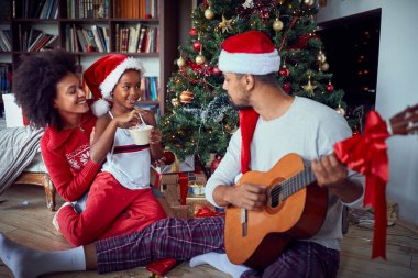  family sitting on floor together and plays on guitar on Christm clipart