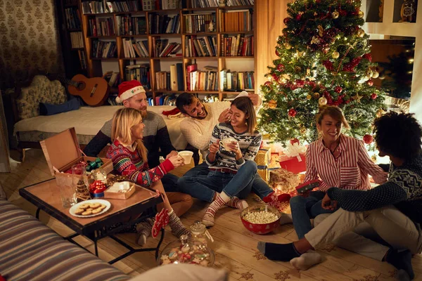 Amigos sentados ao lado de uma árvore de Natal, comer biscoitos de Natal — Fotografia de Stock
