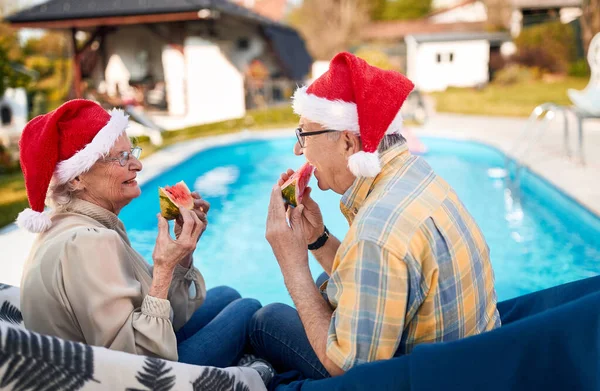Elderly man and woman in Santa hats enjoy on Christmas holiday and eating watermelon — Stock Photo, Image